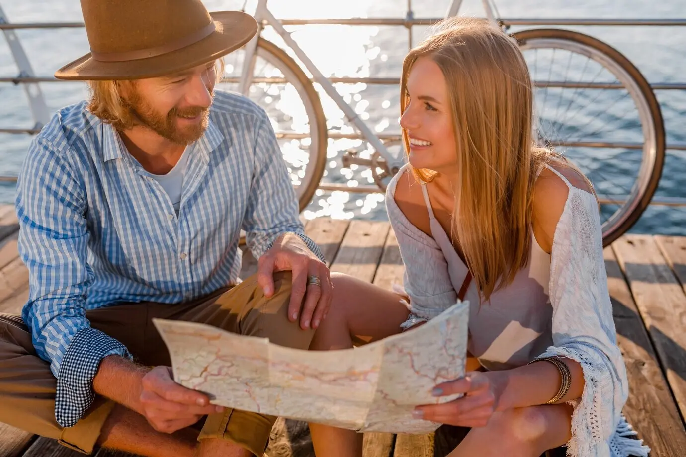 Ein attraktives, fröhlich lächelndes Paar reist im Sommer mit Fahrrädern am Meer; Mann und Frau mit blondem Haar im Boho-Hipster-Stil haben zusammen Spaß und schauen beim Sightseeing auf eine Karte.